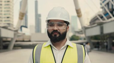 Çağdaş binalara karşı duran miğferli ve gözlüklü genç Hintli inşaatçının portresi. Sakallı endüstriyel yelek uzmanı şehir sokaklarına olan güvenini dile getirdi