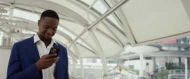 Genç Afrikalı Amerikalı iş adamının portresi iş merkezindeki akıllı telefona bakarken gülümsüyor. Şehirdeki anamorfik lenslerde olduğu gibi cep telefonundaki mutlu ofis çalışanı mesajları