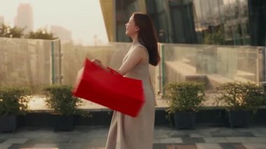 Genç Asyalı kadın portresi alışveriş torbalarıyla iş merkezinin terasında dans ediyor. Güzel Çinli kadın, modern şehir merkezindeki mağaza paketlerini elinde tutuyor.