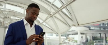 Alışveriş merkezindeki akıllı telefondan Afro-Amerikalı iş adamının portresi. Odaklanmış girişimci ofis lobisindeki anamorfik lenslerde olduğu gibi cep telefonuyla verileri kontrol ediyor