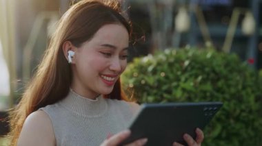Kulaklıklı mutlu Asyalı kadın portresi yeşil parkta dijital tabletle video izliyor. Pozitif Koreli kadın rüzgarlı bir günde şehir merkezindeki mobil cihaz ekranına bakıyor.