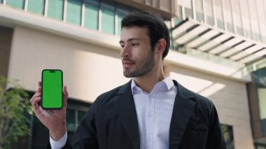 Pozitif İspanyol ofis çalışanının portresi şehir merkezinde renkli ekranlı akıllı telefon gösteriyor. Sakallı Latin girişimci, caddede model görüntülü cep telefonu sunar