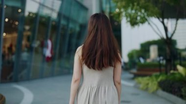 Kolsuz elbiseli genç Asyalı kadın şehrin arka manzarası boyunca yürüyor. Uzun kahverengi saçlı zarif Koreli kadın modern şehir merkezinde tek başına dolaşıyor. Şehir hayatı
