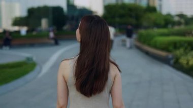 Uzun kahverengi saçlı genç bir kadın şehir parkının arka manzarasında yürüyor. Zarif genç Asyalı bayan modern finans bölgesinde tek başına dolaşırken etrafa bakıyor. Sokak tarzı