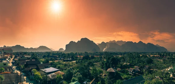 Laos 'ta Vang Viang' ın Smokey Golden Hour Panoraması. Puslu turuncu gökyüzü, öndeki küçük kırsal kasabayla uzak sıradağları boğuyor.