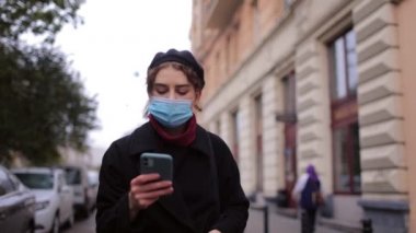 Maskeli kadın sokakta akıllı telefon kullanıyor.