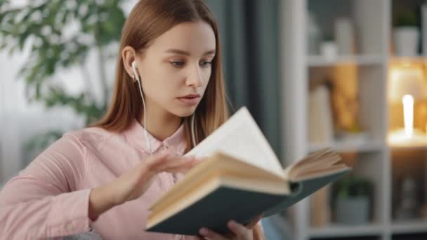 Mulher lendo livro enquanto sentado no sofá em fones de ouvido — Vídeo de Stock