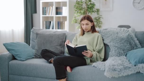 Жінка сидить на дивані і читає книгу — стокове відео