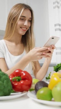 Bir kadın taze sebzelerle dolu parlak bir mutfakta akıllı telefonunu kullanırken gülümsüyor. Telefonuyla meşgul olurken renkli ürünler onu sarıp yemek hazırlıyor..