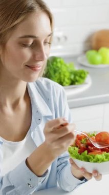 Modern bir mutfakta, bir kadın taze yeşillik ve domatesle renkli bir salata yerken gülümser. Mekan onun çevresindeki doğal ışık ve taze ürünlerle dolu..