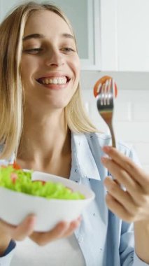 Bir kadın parlak mutfağında taze salata tutarken gülümsüyor. Yeşilliklerden bir ısırık alıyor, sağlıklı yemeğin ve neşeli atmosferin tadını çıkarıyor..