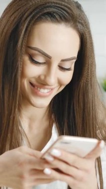 Bir kadın mutfakta, akıllı telefonuna mesaj atarken mutlu bir şekilde gülümsüyor. Sıcak ortam ve ifadesi gün boyunca canlı bir atmosfer yaratıyor..