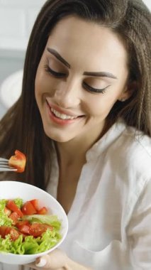 Bir kadın, parlak mutfağında salata yerken gülümsüyor. Taze sebzeler ve domatesler kasesini dolduruyor, sağlıklı ve canlı bir yemek sergiliyor. Beslenmenin keyfini çıkarmak neşe getirir..