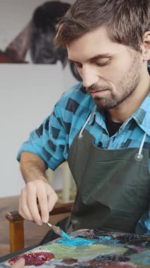 Bir adam stüdyosunda resim yapmaya odaklanmış, paletin renklerini karıştırıyor. Ekose bir gömleğin üzerine önlük giyip fırça darbelerini dikkatlice uyguluyor ve sanatsal yeteneğini yaratıcı bir alanda gösteriyor..