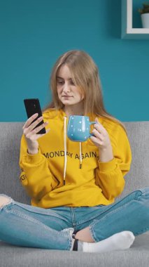 Gri bir kanepede oturan, sarı kapüşonlu bir kadın akıllı telefonuna kaşlarını çatarak bakıyor. Bir elinde mavi bir bardak tutuyor, odaklanmış ve dalgın görünüyor..