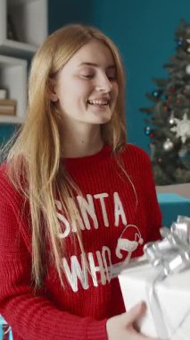 Kırmızı kazaklı genç bir kız güzel paketlenmiş bir hediye kutusunu açarken sevinçle gülümsüyor. Oda tatil için dekore edilmiş, sıcak bir atmosfer yaratıyor..