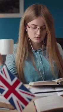 Bir kadın elinde bir fincan çayla evde İngilizce derslerine odaklanıyor. Etrafı kitaplarla ve İngiliz bayrağıyla çevriliyken ses dersleri dinleyip rahat bir çalışma ortamı yaratıyor..