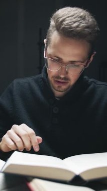 Gözlük takan odaklanmış bir adam karanlık bir alanda kitap okuyor. Yumuşak ışık, sayfaları çevirip anlayışını derinleştirirken yüzündeki ciddi ifadeyi vurgular..