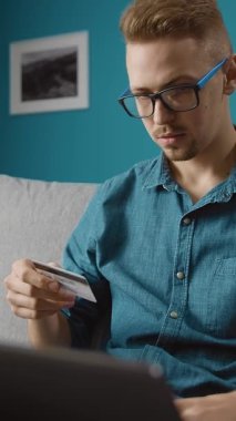 Parlak bir oturma odasındaki koltukta oturan bir kişi elinde kredi kartıyla internette dizüstü bilgisayar arıyor. Satın almaya odaklandıklarında ortam rahatlatıcı ve davetkar..