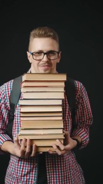 Gözlüklü bir adam siyah arka planda dururken büyük bir yığın kitap tutuyor. Akademik iş yükünü dengelemek için çalışmaya ya da derse katılmaya hazır görünüyor..