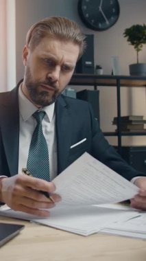 İş kıyafeti giymiş profesyonel bir adam parlak bir iş gününde masasındaki kağıtları incelemeye odaklanır. Ayarda modern ofis dekorasyonu ve organizasyonel unsurlar yer alıyor.