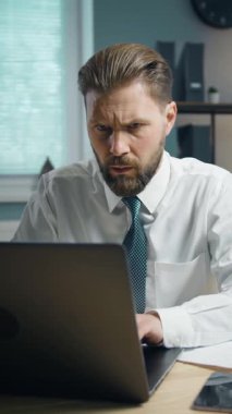 Bir iş adamı iyi aydınlatılmış bir ofiste özenle dizüstü bilgisayarının başında çalışır. İş saatlerinde görevleri yönetirken ve etkili bir şekilde iletişim kurarken ifadesi konsantrasyonunu gösteriyor..