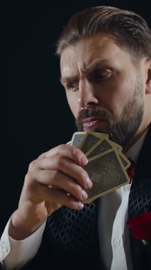 Loş bir odada, şık giyinmiş bir adam zarif kartlar tutarken dikkatle odaklanır. Duruşu ve şık kıyafeti entrika ve sofistike bir atmosfer yaratıyor..