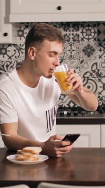 Bir adam sabah kahvaltısı sırasında meyve suyunu yudumlayarak masada oturur ve akıllı telefonuna bakar. Tabağında tost var..