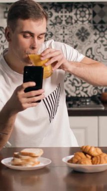 Bir adam sabah yemeğini evde yer, akıllı telefonunu kontrol ederken meyve suyunu yudumlar ve taze ekmek ve hamur işlerinin tadını çıkarır..