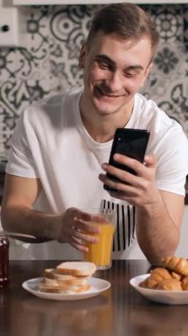 Genç bir adam kahvaltı masasında oturuyor, akıllı telefonuna bakıyor ve parlak bir mutfakta meyve suyu içiyor..