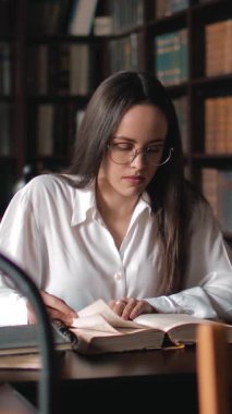 Bir kadın kütüphanede bir masada oturmuş, açık bir kitaptan okumaya ve not almaya odaklanmış..