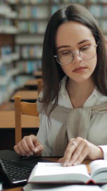 Bir öğrenci, kitaplarla ve notlarla çevrili bir kütüphane masasında derslerine odaklanır. Öğrenmek için huzurlu bir atmosferin tadını çıkarır..