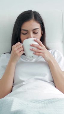 Yatakta dinlenen biri, sabah güneşinin dinginliğini kucaklarken sıcak bir fincan kahvenin tadını çıkarır..