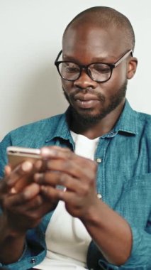 Siyah bir adam evde otururken akıllı telefonunu kontrol eder. Odaklanmış ve ekranla meşgul görünüyor..