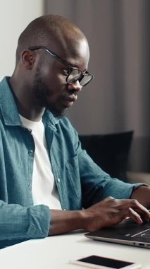 Siyah bir adam, modern bir ortamda basit bir masada dizüstü bilgisayarı üzerinde çalışıyor..