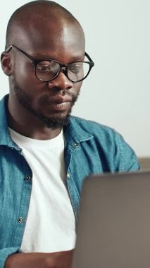 Siyah bir adam, kapalı bir alanda dizüstü bilgisayarı üzerinde çalışıyor. Meşgul ve işine odaklanmış görünüyor..