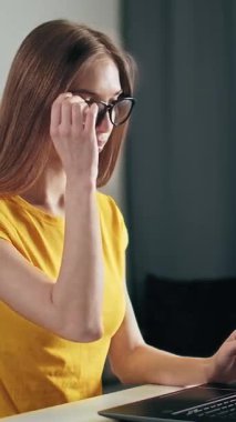 Bir kadın, dizüstü bilgisayarına odaklanmış bir şekilde masada oturur ve gözlüğünü sakin bir ortamda düşünceli bir şekilde ayarlar..