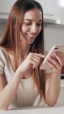 Bir kadın, modern bir mutfakta oturmuş, zamanını geçirirken akıllı telefonuyla gülümsüyor..