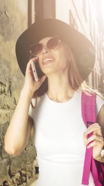 Sırt çantalı ve şapkalı bir turist, güneşli bir sokakta telefonla konuşurken gülümsüyor..