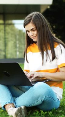 Güneşli bir parkta çimlerin üzerinde oturan bir kadın konsantre bir şekilde çalışırken dizüstü bilgisayarına odaklanıyor.