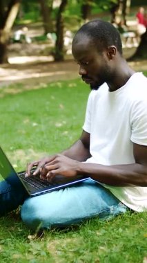 Parktaki çimlerin üzerinde oturan bir adam güneşli bir günün tadını çıkarırken dizüstü bilgisayarına odaklanıyor.