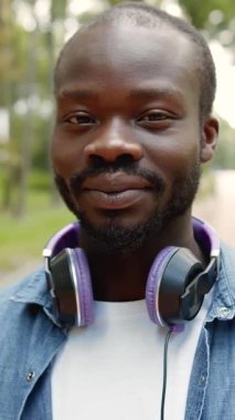 Güneşli bir günde parkta yürürken kulaklık takan neşeli bir adam gülümsüyor.