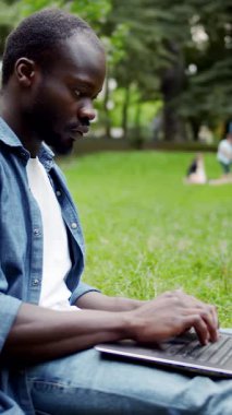 Parktaki çimenlerde oturan bir adam dışarıda manzaranın tadını çıkarırken dizüstü bilgisayarıyla mutlu bir şekilde çalışıyor.