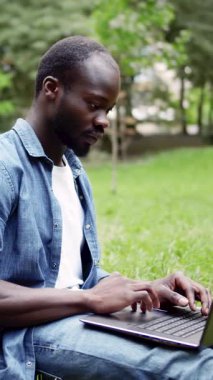 Parkta dizüstü bilgisayarı üzerinde çalışan bir adam, yazarken rahatlatıcı yeşil ortamın ve temiz havanın tadını çıkarıyor.