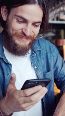 Sosyal medya ve dijital bağlantılara dalmış rahat bir kafede akıllı telefonunu kullanan bir adam gülümsüyor.