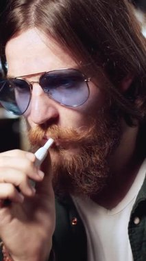 Sakallı ve güneş gözlüklü bir adam, gevşemiş bir atmosferle çevrili bir şekilde kafasına bir kurşun yiyip öksürüyor.