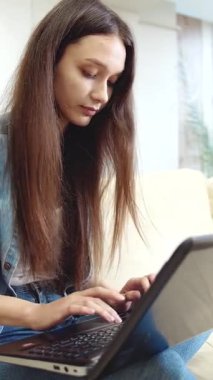 Bir kadın rahat evinde dizüstü bilgisayarını kullanarak kanepesinde oturuyor.