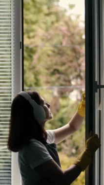 Bir kadın gün ışığında pencereyi sarı eldivenle temizlerken kulaklık takıyor..