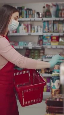 Maskeli bir pazar çalışanı elinde kırmızı bir sepet tutarken raftan eşyalar seçer.