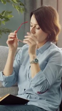 Kadın oturma odasındaki kanepede kitap okurken gözlük takıyor..